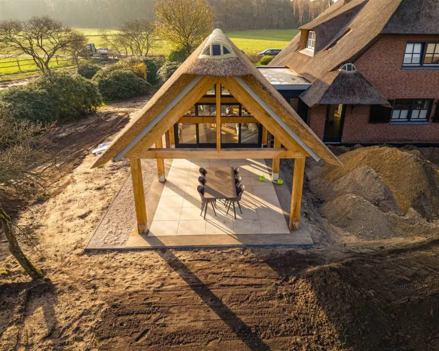 Een houten veranda met rieten kap in aanbouw, tegen een woning met rieten dak