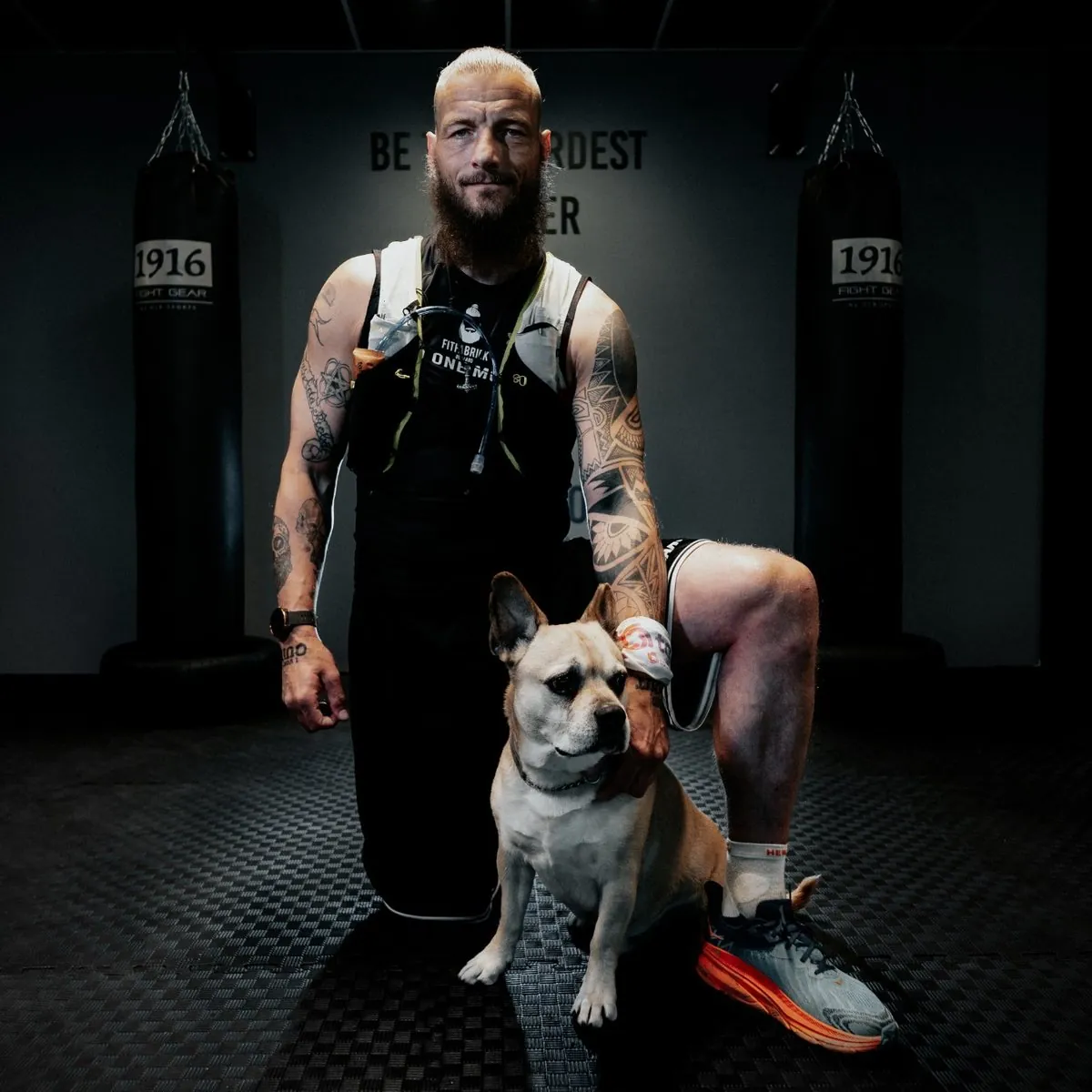 Ralf van Fitfabriek de Baard, geknield met zijn hond tussen de bokszakken in de sportschool